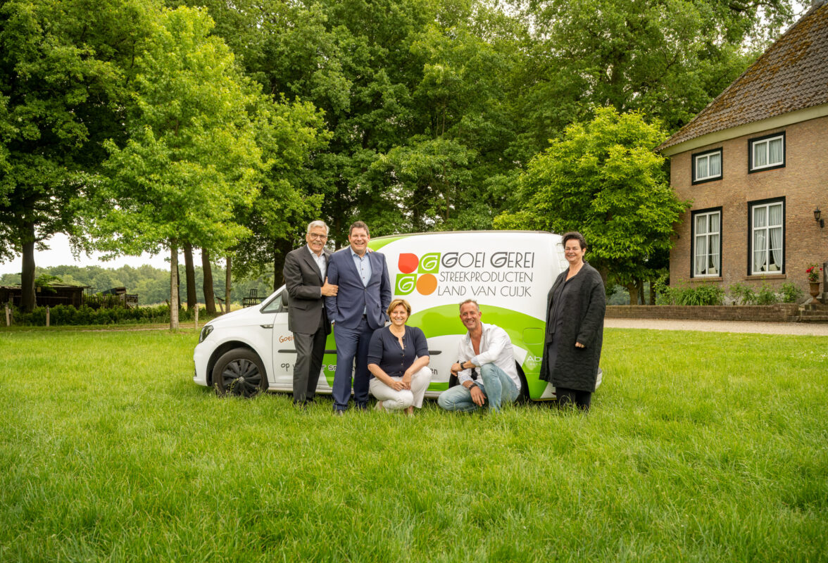 Vijf mensen poseren bij een bestelbus met het logo ‘Goei Gerei Streekproducten Land van Cuijk’, naast een boerderij en groene bomen.